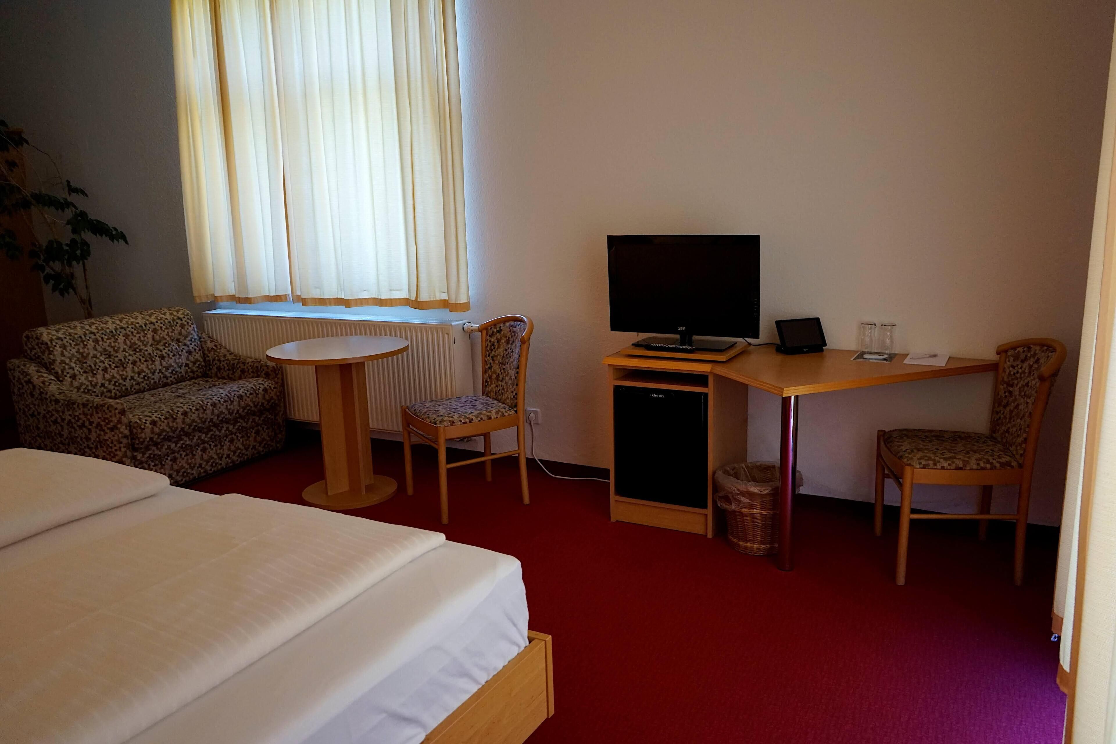 A hotel room with a bed, a desk with a computer, a chair, a sofa, and a window with curtains.