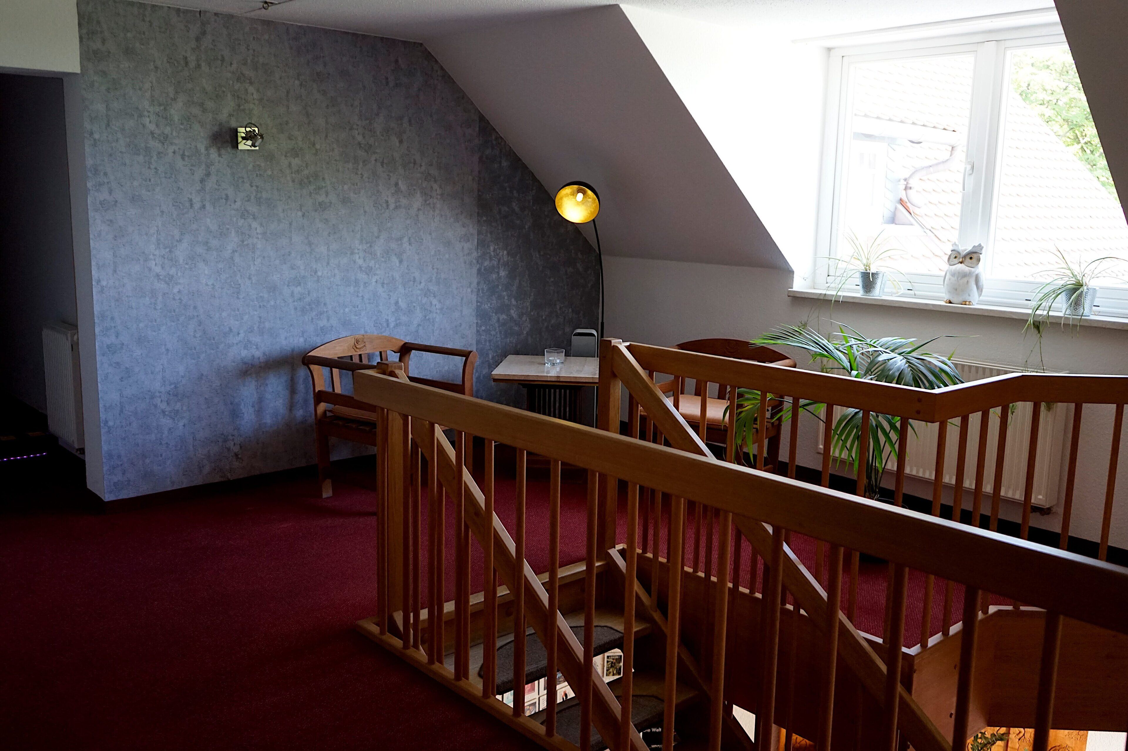 A room with a sloped ceiling, a wooden staircase, a red carpet, and a window with plants on the sill.