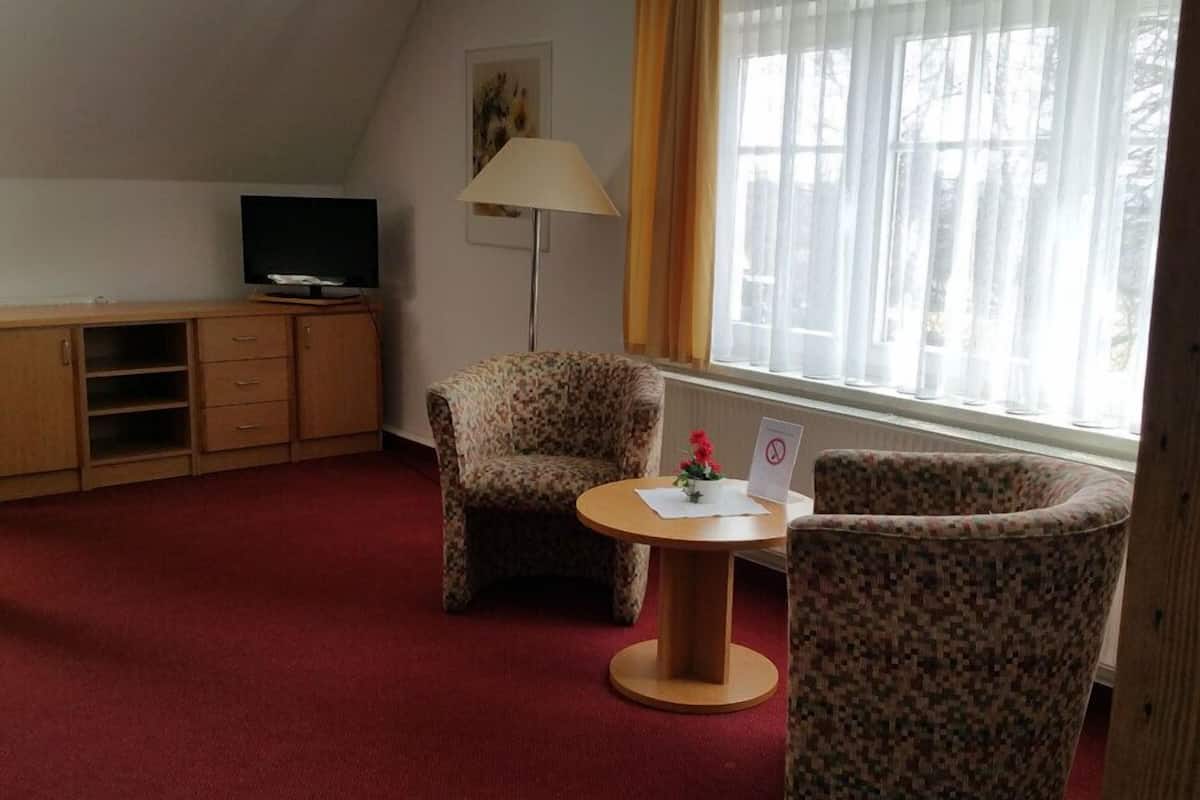 Ein Hotelzimmer mit einem Holztisch, einem Fernseher, einem blumigen Ohrensessel, einem runden Tisch mit Blumenvase und einem Fenster mit Vorhängen.