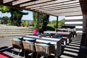 Eine Terrasse im Freien mit Holztischen und Korbstühlen, umgeben von Topfpflanzen und Blick auf die Landschaft.