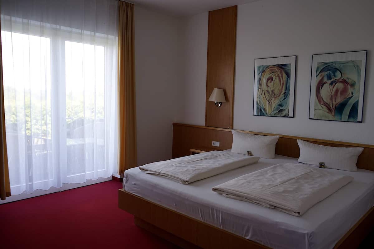 Ein Hotelzimmer mit einem großen Bett, einem Holzkopfstück, zwei Bildern an der Wand, einer Lampe und Blick durch transparente Vorhänge.
