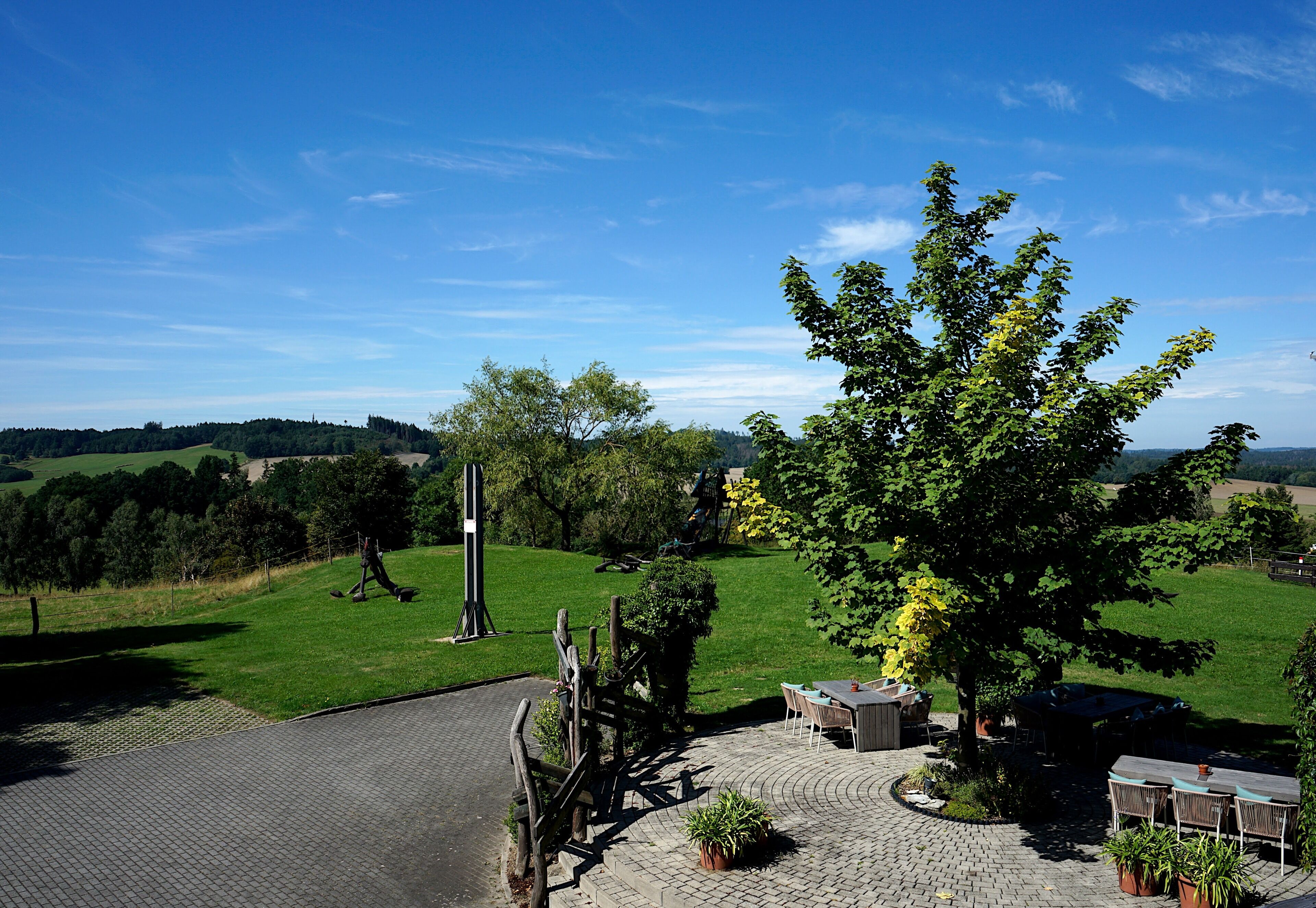 Ein gepflasterter Patio-Bereich mit Gartenmöbeln, ein großer Baum und Blick auf ein Wiesfeld und ferne Bäume.