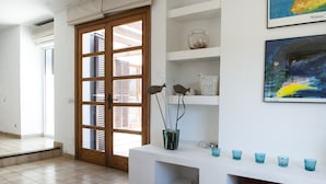 A room with a white tiled floor, a wooden door with glass panels, a white console table with blue glass cups, a wicker basket, and a vase with branches.
