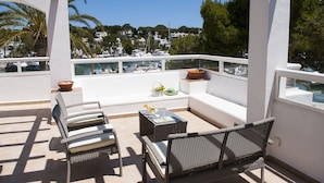 A balcony with wicker furniture, a small table with a vase, and a view of boats and houses in the background.
