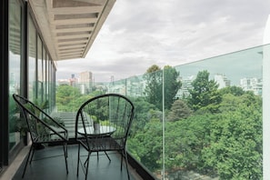 Un balcón con dos sillas de mimbre, una mesita y baranda de cristal con vistas a un paisaje urbano y zonas verdes.