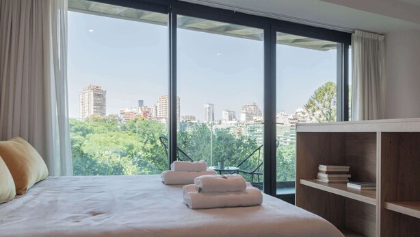 A hotel room with a large bed, a wooden nightstand, and a view of the city through large windows. - MOT Rentals Armenia Luxury Lofts Mansion Botanico (Palermo Soho)