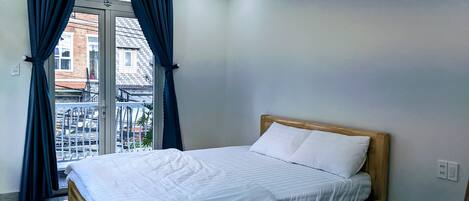 Un dormitorio con cama, ropa de cama blanca y vistas a edificios a través de una ventana con cortinas azules.