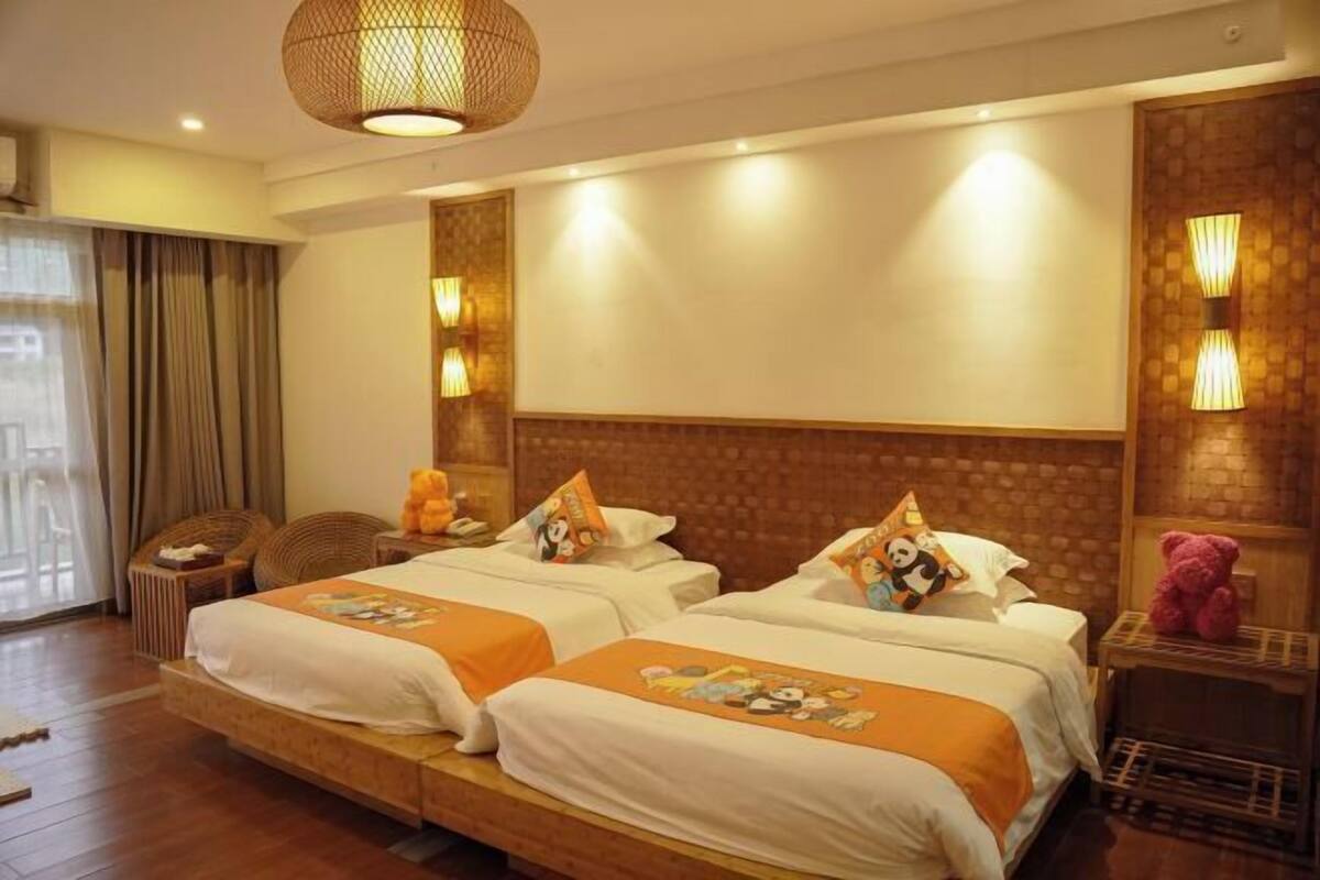 A hotel room with two beds, a large window with curtains, a wicker chair, and a small table with a teddy bear.