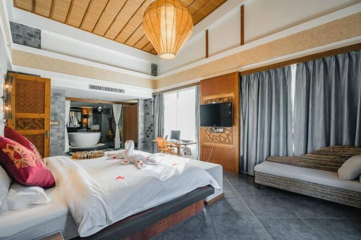 A spacious bedroom with a large bed, a sofa, a TV, and a bathroom visible through an open door.