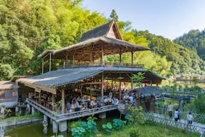 A traditional wooden pavilion with a thatched roof, surrounded by lush greenery and a pond.