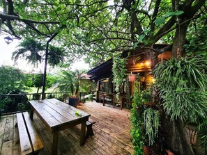 Une terrasse extérieure en bois, avec une table et des bancs en bois, entourée d’une végétation luxuriante et d’arbres.