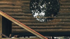 A rustic wooden structure with a circular mirror reflecting trees, a wooden sloped roof, and a stack of logs.