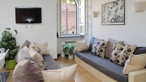 A living room with a grey sofa, multiple cushions, a flat-screen TV, a potted plant, and a window with a view of a brick wall. - Designer Duplex 2BR 2BA by Colosseum - Historic Center, Metro (Roma)