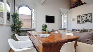 A dining area with a wooden table, white chairs, a television, and a large window with a view of a brick wall. - Designer Duplex 2BR 2BA by Colosseum - Historic Center, Metro (Roma)
