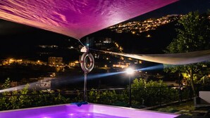 Uma banheira de hidromassagem com interior iluminado em roxo, rodeada por um toldo escuro, com vista para um horizonte urbano à noite.