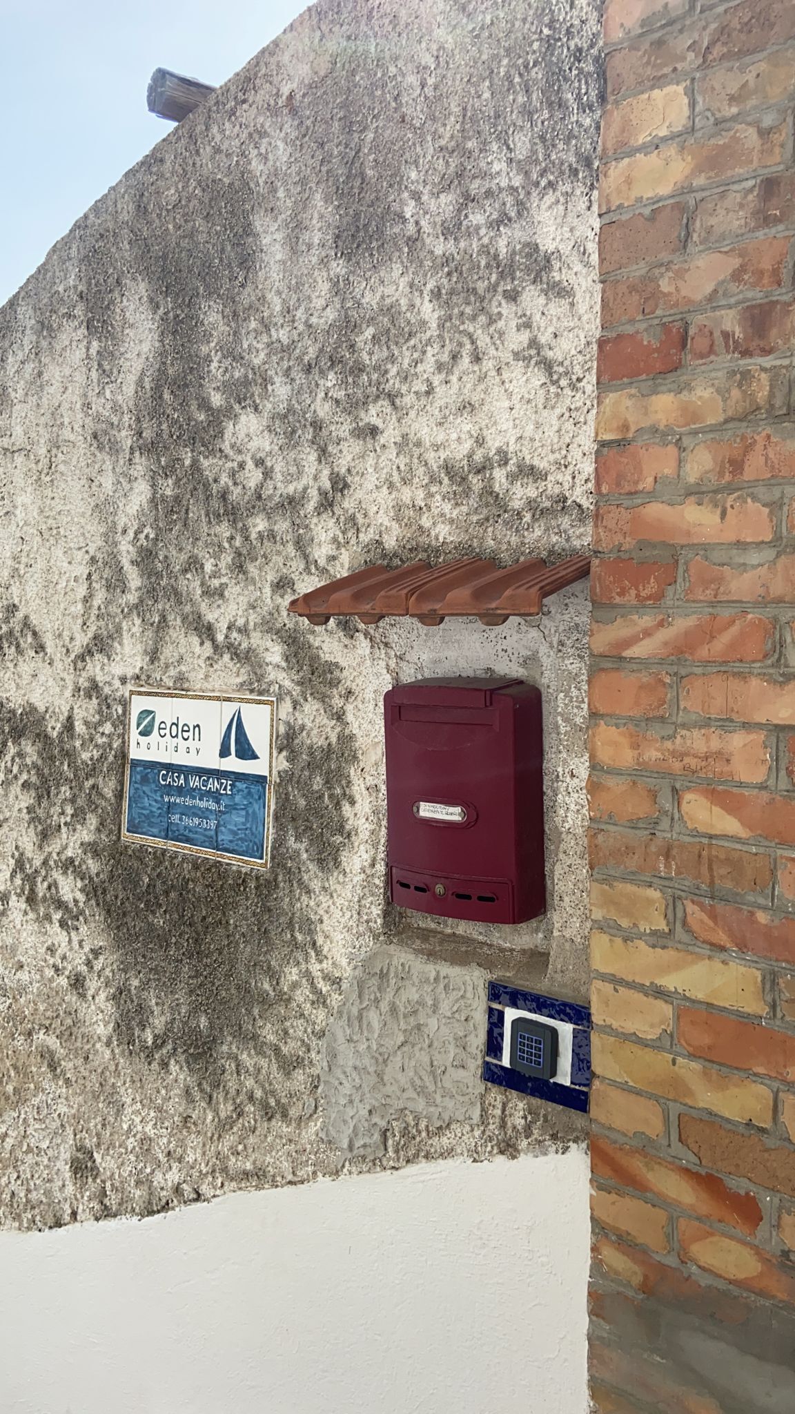 A sign for 'eden holiday' indicating a vacation rental property, a red electrical box, and a blue electrical meter on a textured wall.