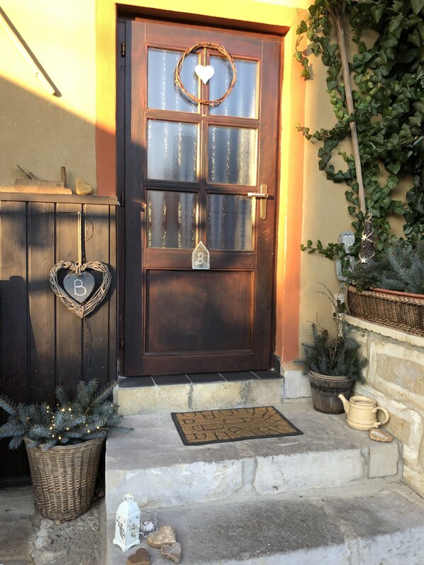Une porte en bois ornée d’une guirlande en forme de cœur et d’une enseigne avec la lettre « B », un sapin de Noël en pot et un banc en pierre avec un paillasson.