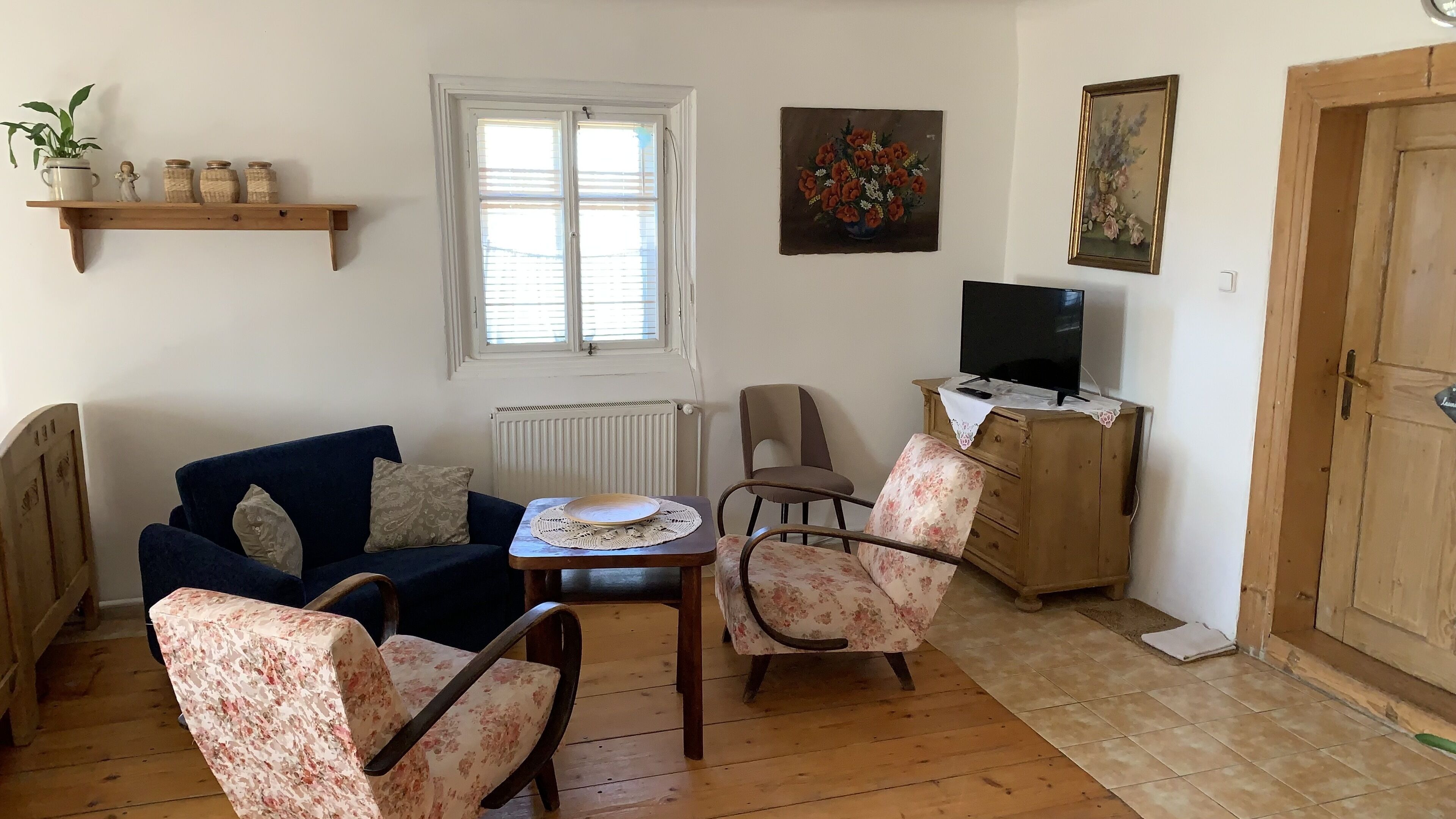 A cozy living room with a blue sofa, floral armchairs, a small table, a TV on a wooden cabinet, and a window with blinds.
