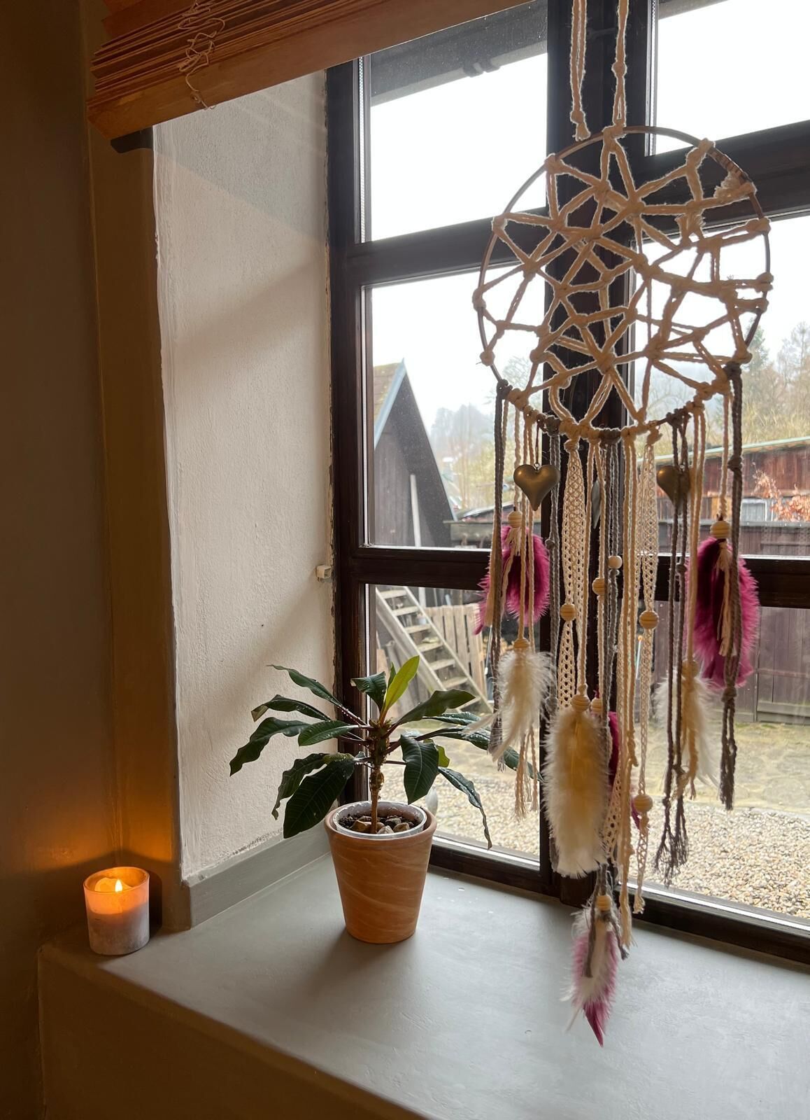 A dreamcatcher hanging by a window with a potted plant on the sill.