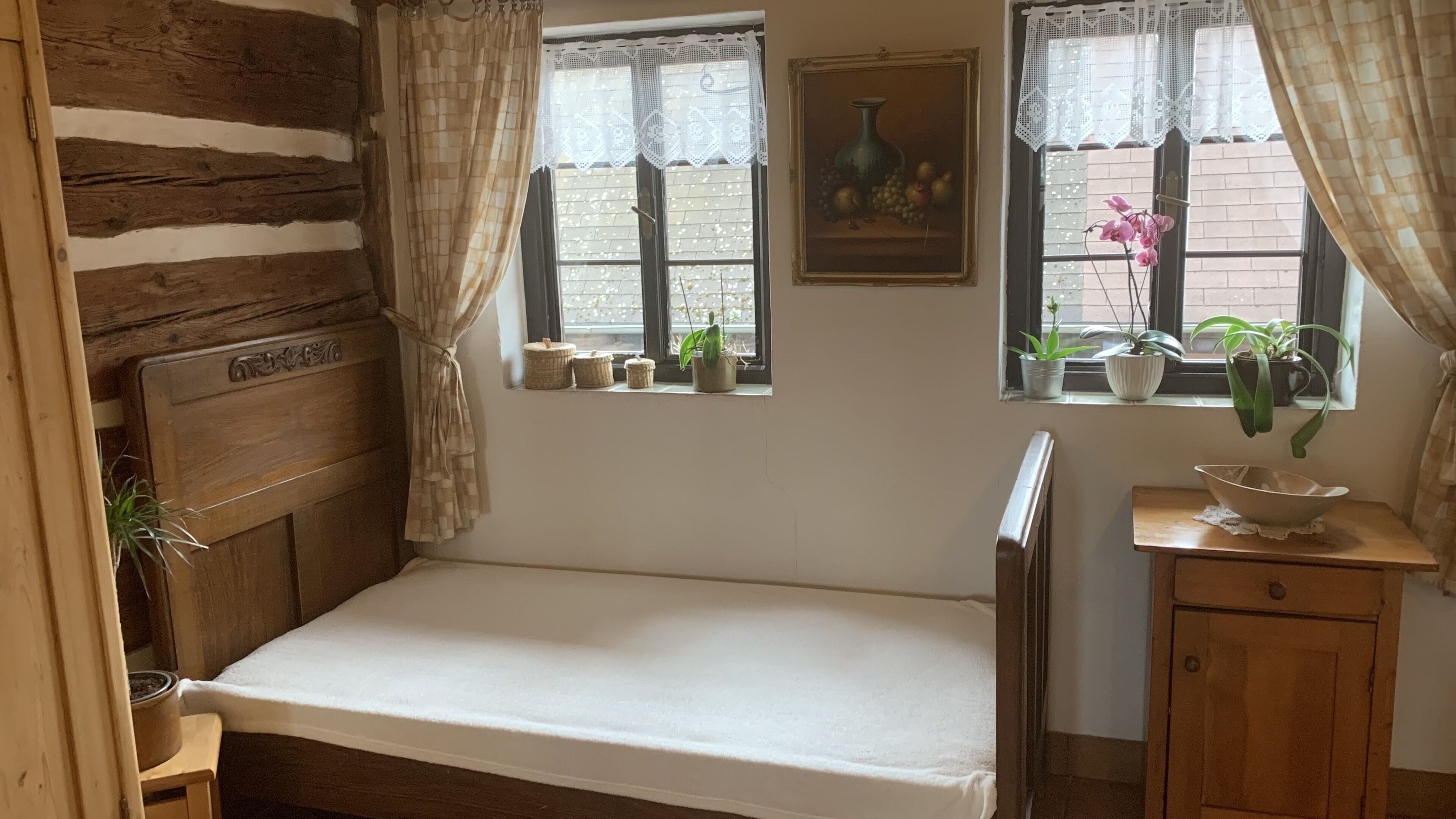 A small, cozy room with a wooden headboard bed, two windows with lace curtains, a small table with a bowl, and potted plants.