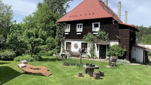 A two-story house with a red roof, a front porch with a bench, and a garden with a lawn and trees. - Pension Laura (Chribska)