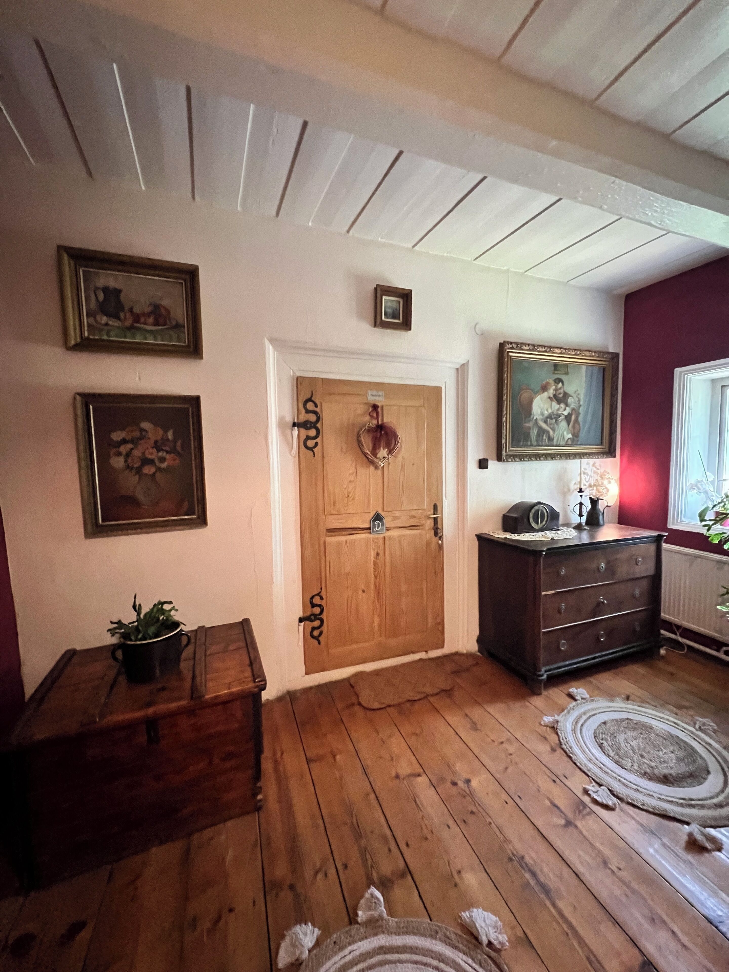 A wooden-floored hallway with a wooden door, a dresser, and framed artwork on the walls.
