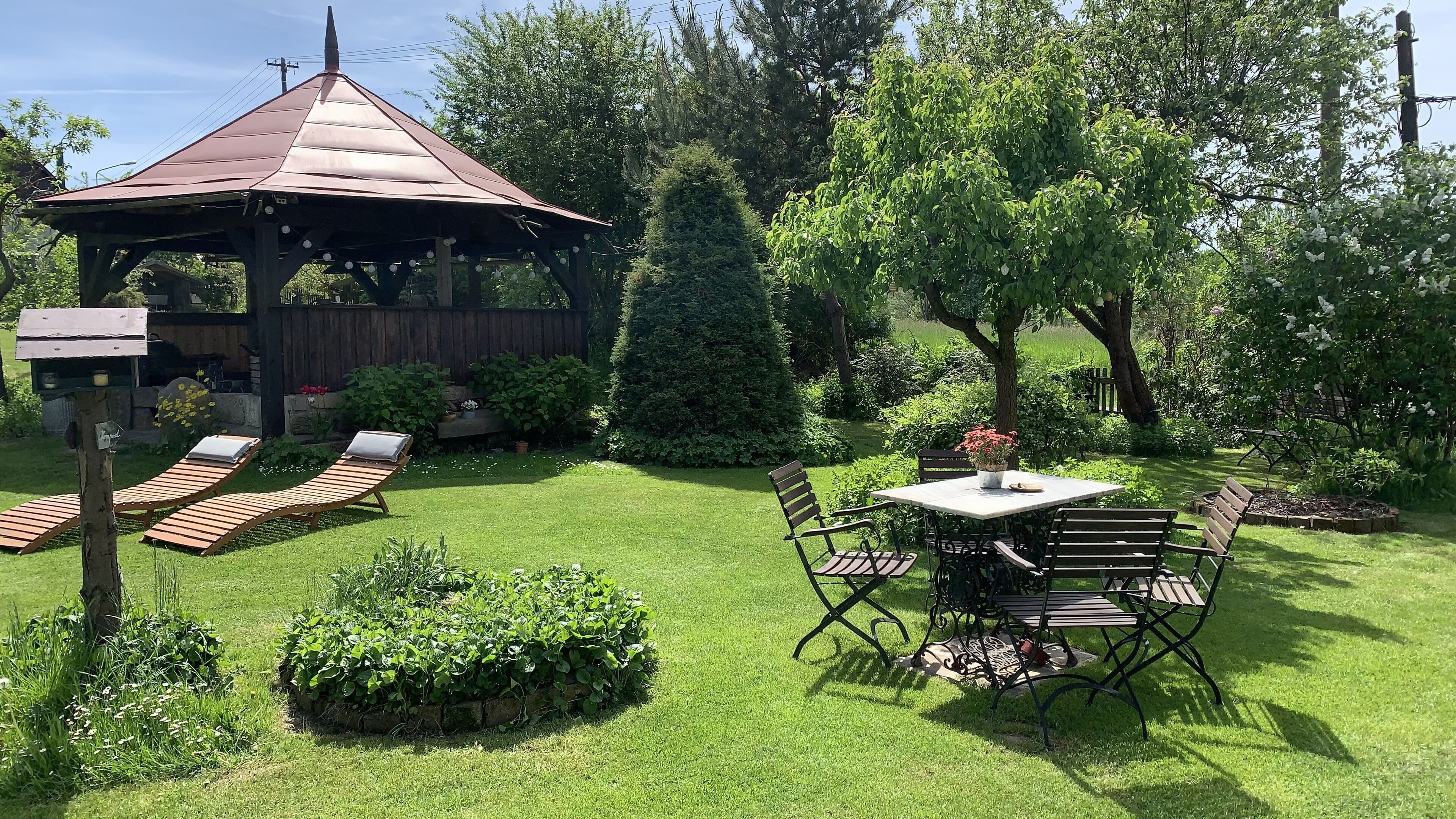A gazebo with a sloped roof, surrounded by a well-maintained lawn and various plants.