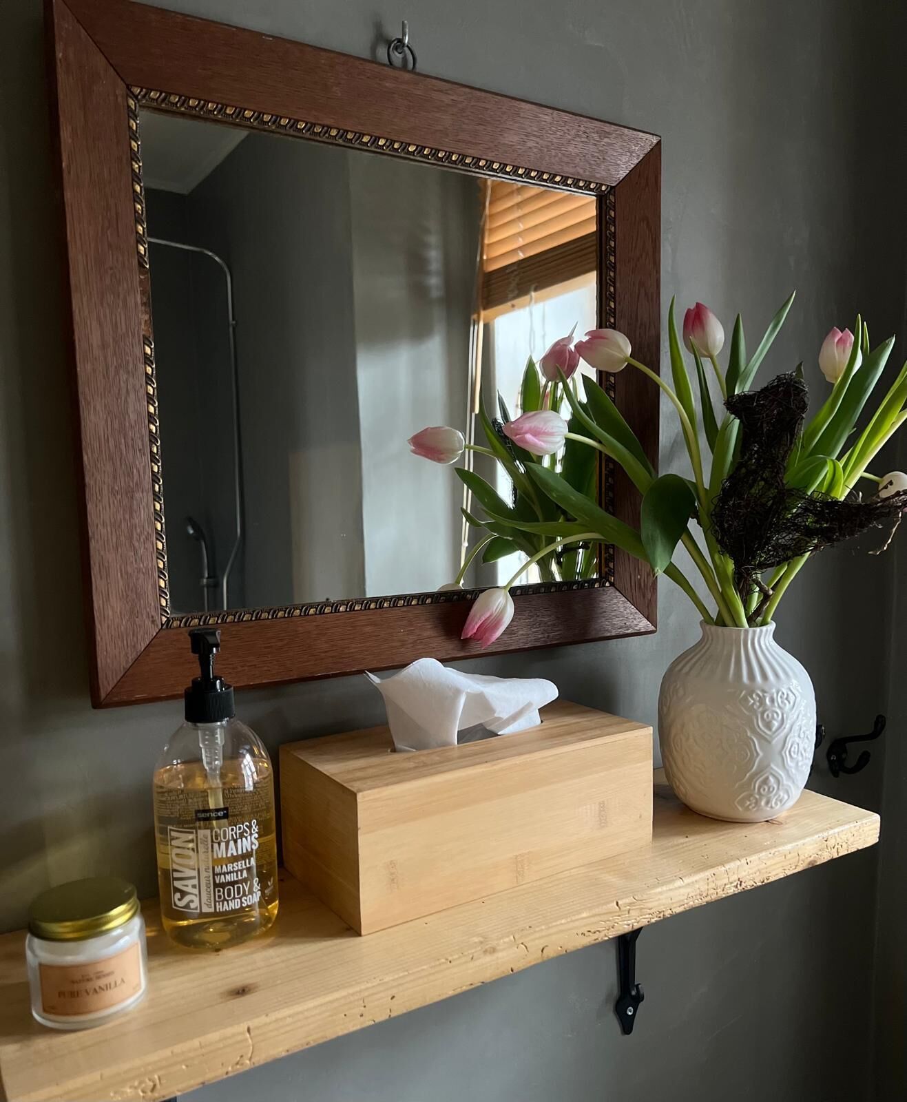 Een houten wastafel met spiegel, een vaas met bloemen, een zeepdispenser en een zakdoosje.