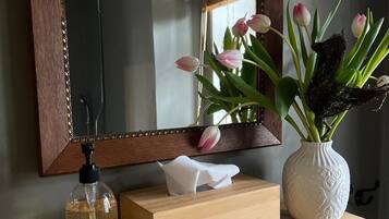 Een houten wastafel met spiegel, een vaas met bloemen, een zeepdispenser en een zakdoosje.