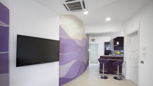 A modern hotel lobby with a flat-screen TV mounted on the wall, a purple and white decorative wall, and a bar area with two purple stools. - Estate4home- L'Onda Viola (Sorrento)