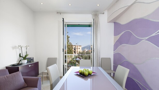 A modern room with a dining table, chairs, a sofa, and a balcony with a view of buildings and mountains. - Estate4home- L'Onda Viola (Sorrento)