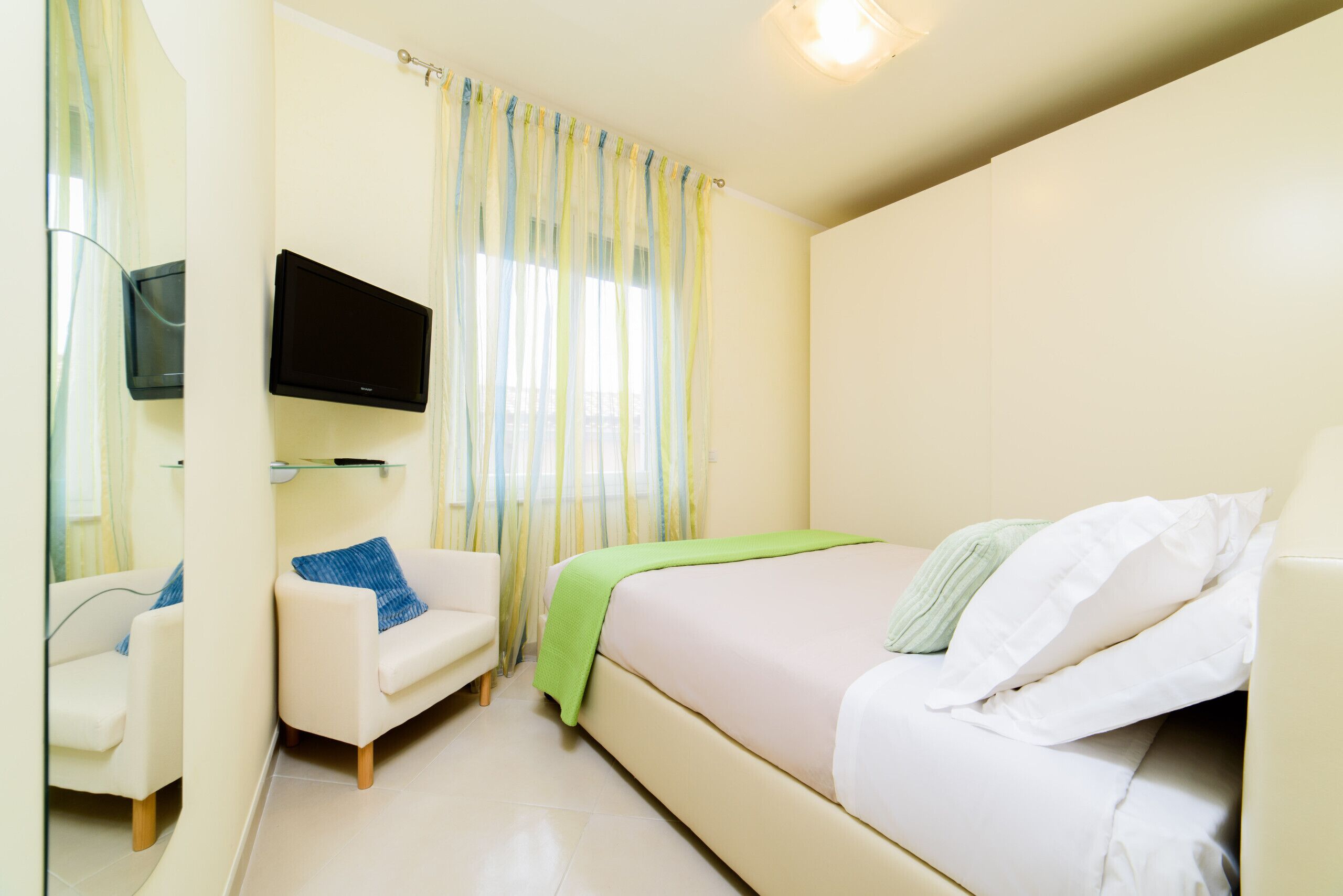 Habitación de hotel con cama, sillón, televisor montado en la pared y ventana con cortinas.