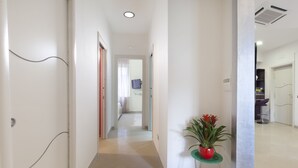 A clean, well-lit hallway with a potted plant, leading to a room with a bed and a bathroom. - Estate4home- L'Onda Viola (Sorrento)