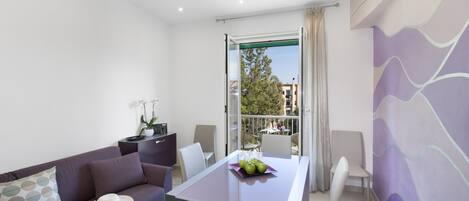 Una sala de estar moderna con un sofá, una mesa de comedor con sillas y un balcón con vistas a edificios y árboles.