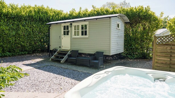 A small, single-story cabin with a door and windows, a hot tub in the foreground, and a wooden fence in the background. - Shepherd's hut with hot tub in its own secluded garden (Longtown)