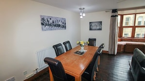 Uma sala de jantar com mesa de madeira, cadeiras pretas e um vaso de flores. Há duas fotografias a preto e branco nas paredes.
