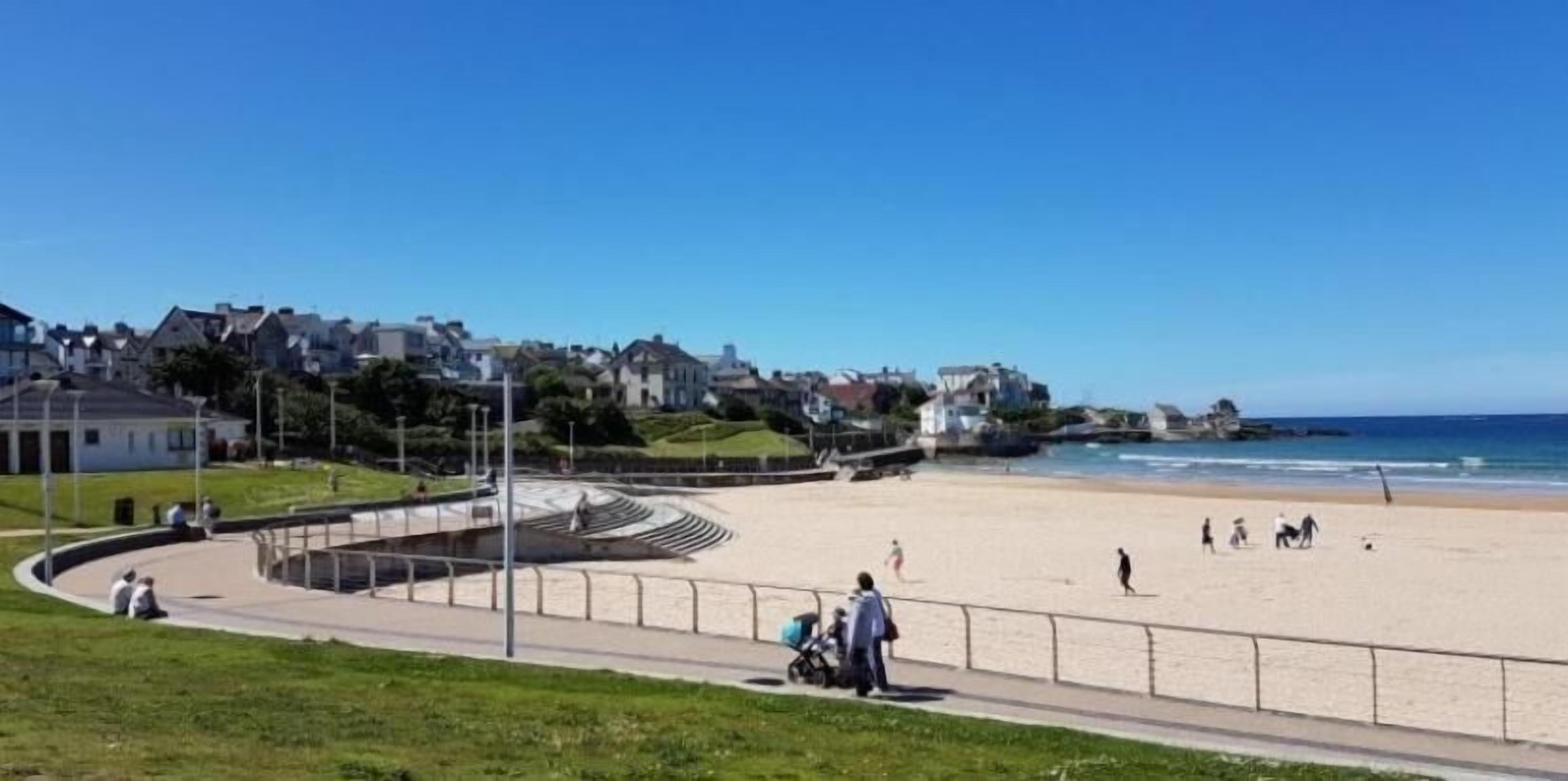 Ein Strand mit einer gewundenen Promenade, Menschen unterwegs und Häuser im Hintergrund.
