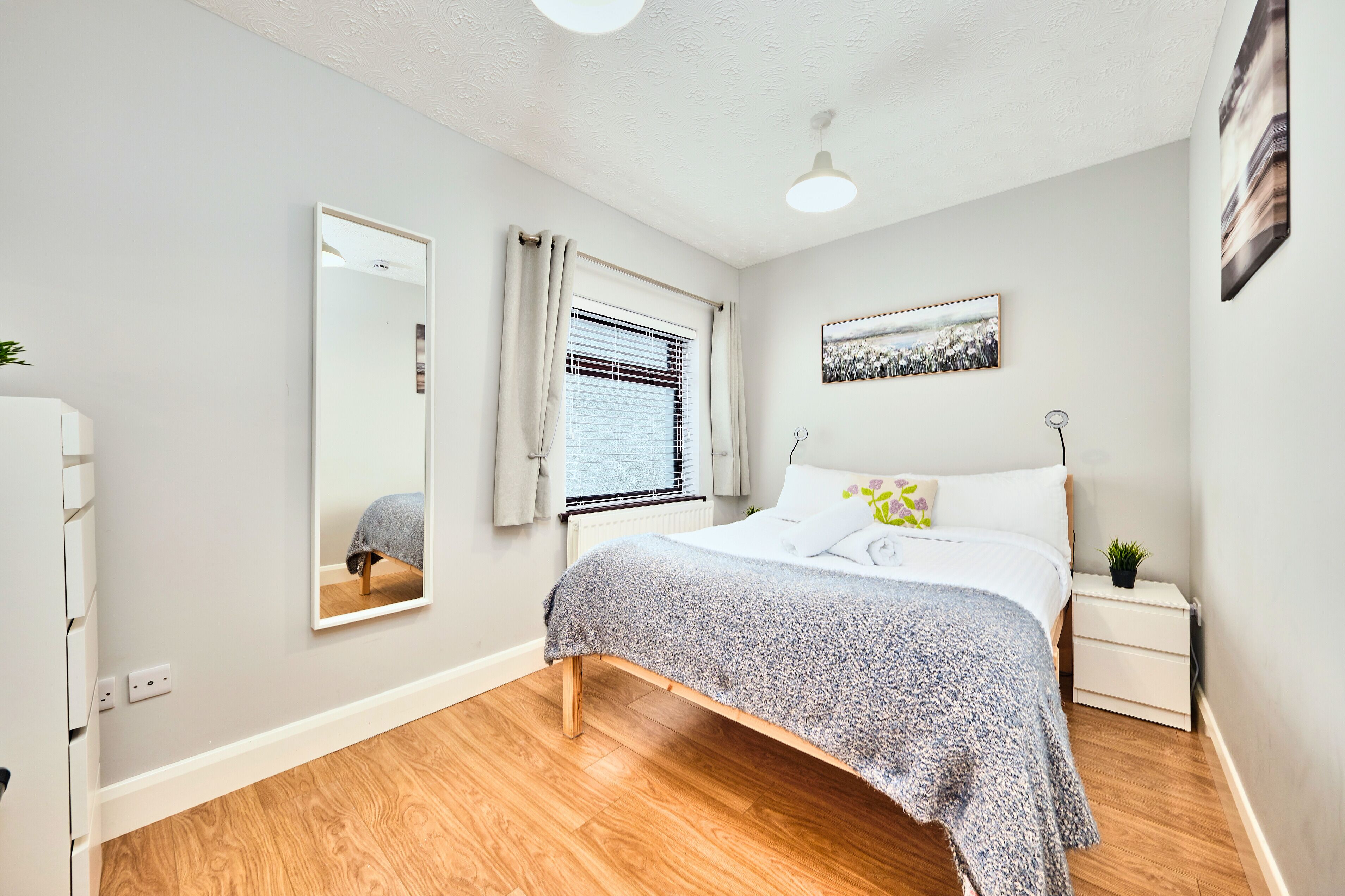 A bedroom with a bed, a bedside table, a nightstand, a mirror, a window with curtains, and a wall-mounted picture.