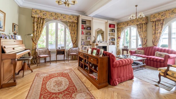 Ampla sala de estar com um piano de cauda, um sofá vermelho e uma mesa de centro. Cortinas florais e uma janela grande proporcionam iluminação natural.