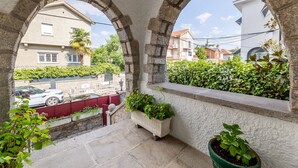 Uma varanda com arcos de pedra, plantas em vasos e vista para a rua com casas e um carro estacionado.