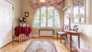 Um quarto com uma grande janela em arco, um radiador, uma mesa com toalha vermelha, uma cadeira, um abajur e um tapete floral.