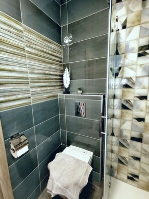 A bathroom with dark tiled walls, a shower area with a glass door, a towel rack, and a small potted plant.