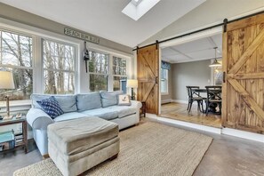 A spacious living room with a light blue sofa, a beige ottoman, and a large window overlooking a tree-lined street. - Steps to Dock Square & Gooch's Beach! Pets Welcome! (Kennebunk)