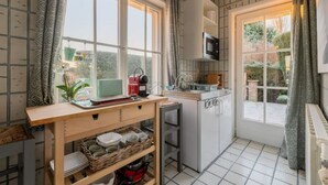 Une cuisine compacte équipée d’un four à micro-ondes, d’un évier et d’un petit réfrigérateur. On y trouve une table en bois avec de la vaisselle et un panier rempli de bols. La cuisine offre une vue sur l’extérieur grâce à une fenêtre agrémentée de rideaux.