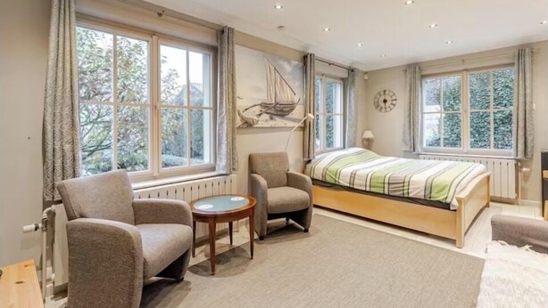 Une chambre à coucher comprenant un lit, deux fauteuils, une petite table et de grandes fenêtres avec des rideaux.