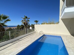 Une piscine sur le toit offrant une vue dégagée sur l’océan, entourée de palmiers et bordée d’un bâtiment blanc.