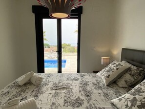 Een slaapkamer met een bed, kussens, een nachtkastje, een lamp en uitzicht op een zwembad en gebouw door een glazen deur.