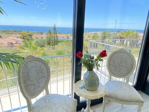 A balcony with two chairs, a small table with a vase of flowers, and a view of the ocean and landscape.