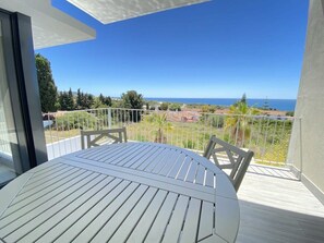 A balcony with a white table and chairs, offering a view of the sea and surrounding greenery.