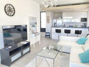 A modern living room with a flat-screen TV, a glass coffee table, a white sofa with blue cushions, and a kitchen in the background.